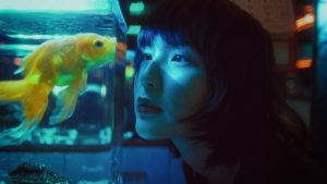 Side view of curious Asian female looking at goldfish swimming in aquarium in dark room