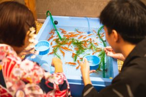 Back view anonymous teenage brother and sister using scoop net to catch small orange freshwater fish from basin to put in fishbowl